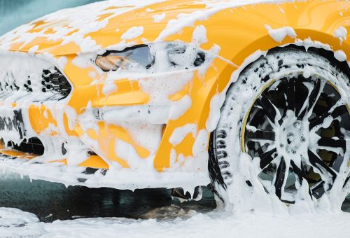 Cropped image of wheel of luxury yellow car in outdoors self-service car wash, covered with cleaning soap foam.; Shutterstock ID 2143537941; purchase_order: -; job: -; client: -; other: -